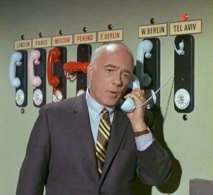 Man in vintage suit making call on old-fashioned wall phones labeled with nostalgic city names from a classic photo.