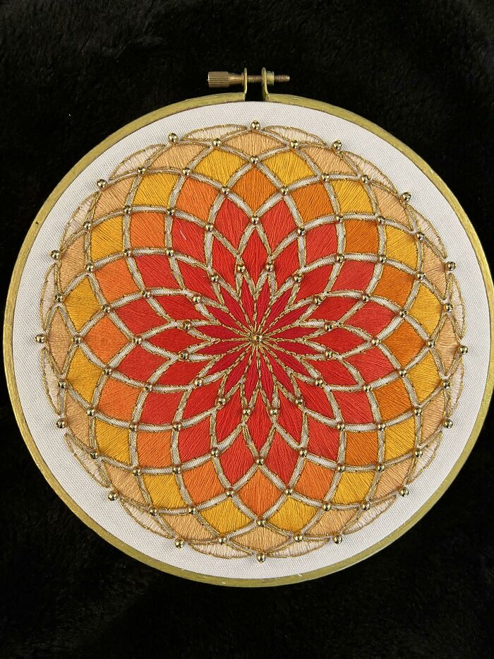 Intricate circular embroidery design with warm orange and red shades accented by gold thread and beads in a hoop frame.
