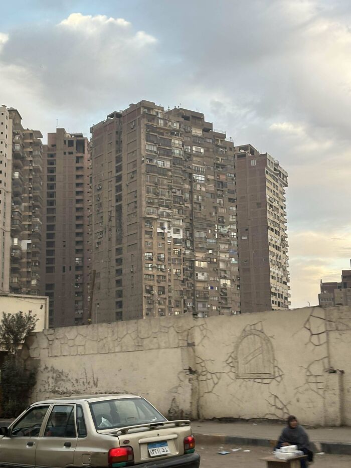 Urban Hell: Deteriorating high-rise buildings under cloudy skies, with a parked car and graffiti-decorated wall in foreground.