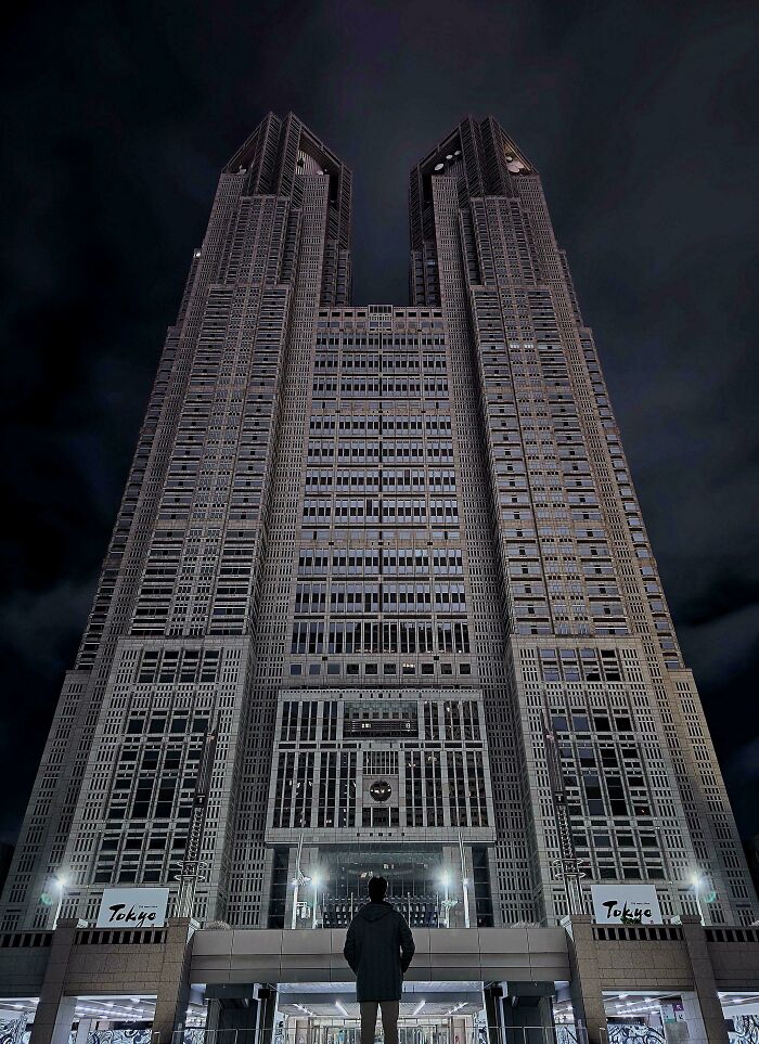 Man standing in front of a towering building with an evil aura, resembling a supervillain headquarters at night in Tokyo.