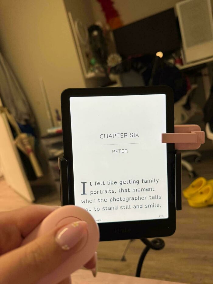 Confusing perspective of a hand holding an open Kindle, displaying Chapter Six text, in a cluttered room.