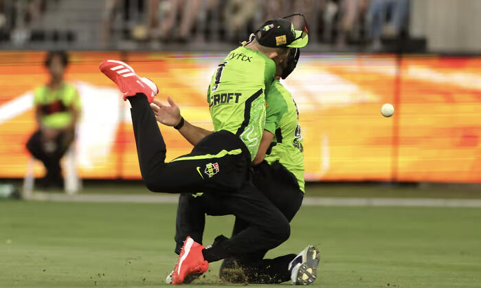 Two cricketers in green uniforms appear to collide on the field, creating a confusing perspective.