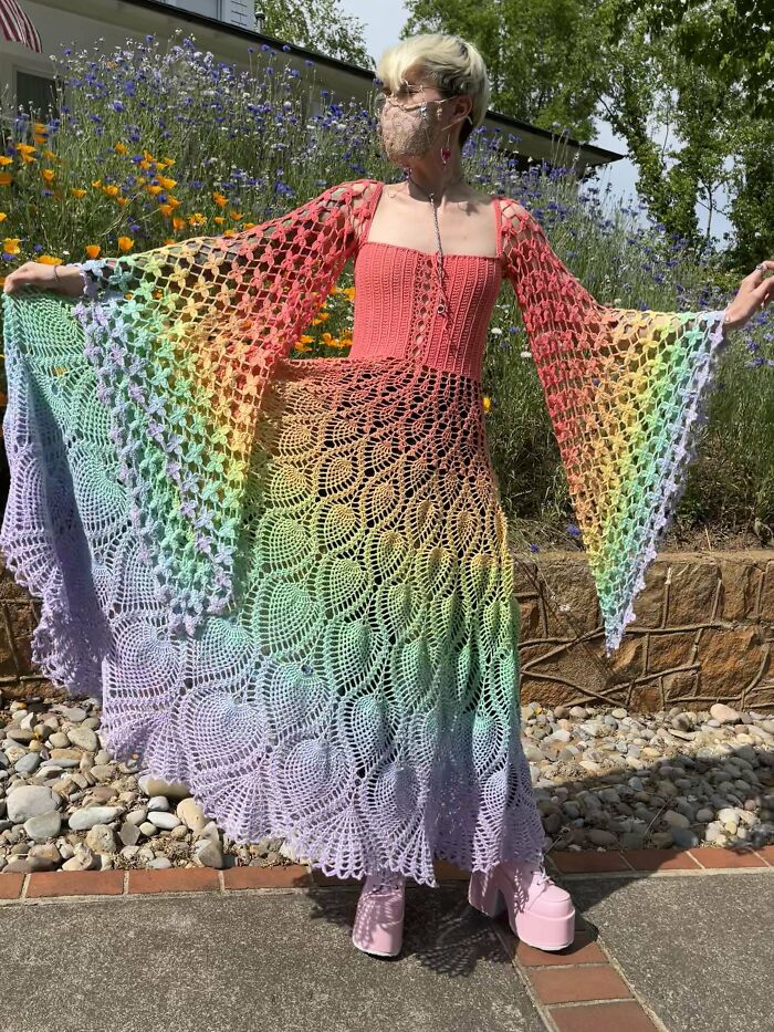 Person wearing colorful crocheted dress with intricate patterns, standing outdoors in a garden.