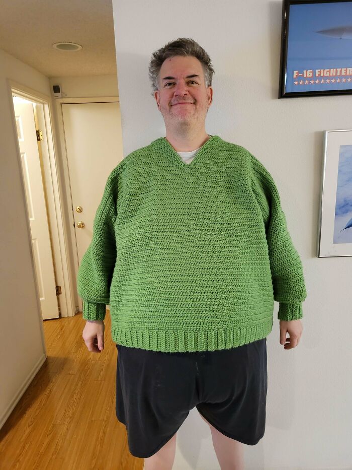 Man wearing a green crocheted sweater, standing indoors, showcasing homemade knitting creativity.