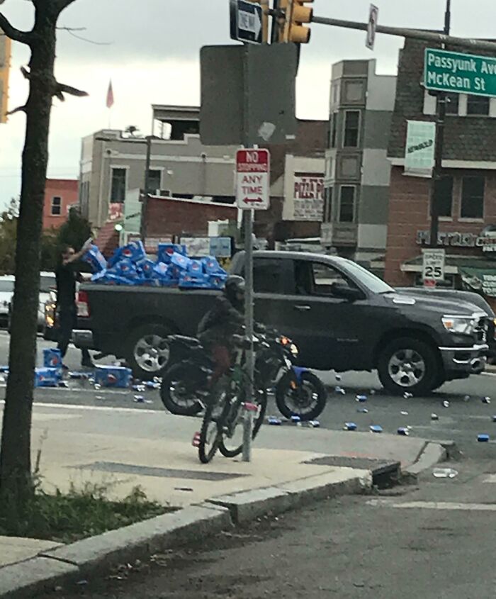 Truck spilling soda boxes at an intersection, man and motorcycle nearby; scene from Idiots-In-Cars series.
