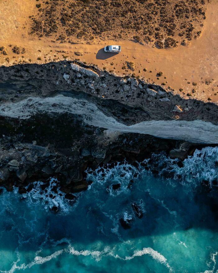 Aerial view of Australian coastline with a camper van on the cliff edge, overlooking the ocean waves below.