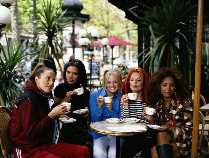 Five women in colorful '90s outfits sitting at a café table, sipping tea, with plants in the background.
