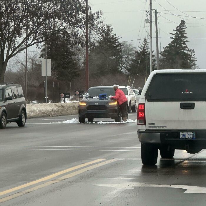 Person clearing snow off a car in the middle of the road, illustrating careless driving.