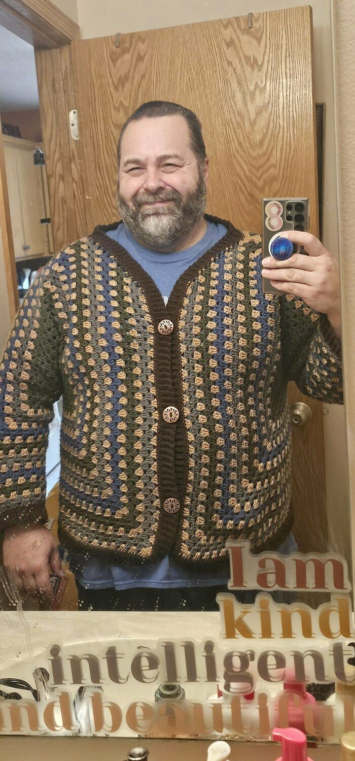 Man smiling and wearing a handmade crochet sweater with buttons taking a mirror selfie showing proud crochet project.