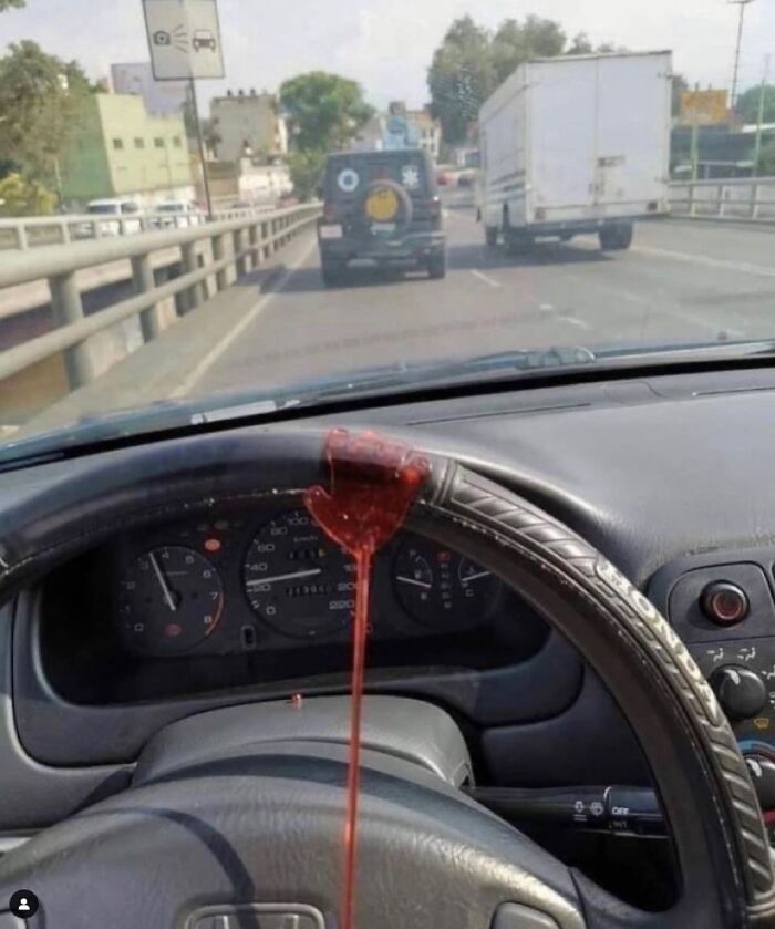 Steering wheel inside car with a thick red liquid resembling blood dripping from the top on a busy road.