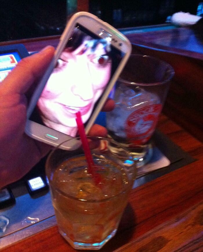 Funny camera roll find with a phone showing a face and a drink with a straw at a bar.