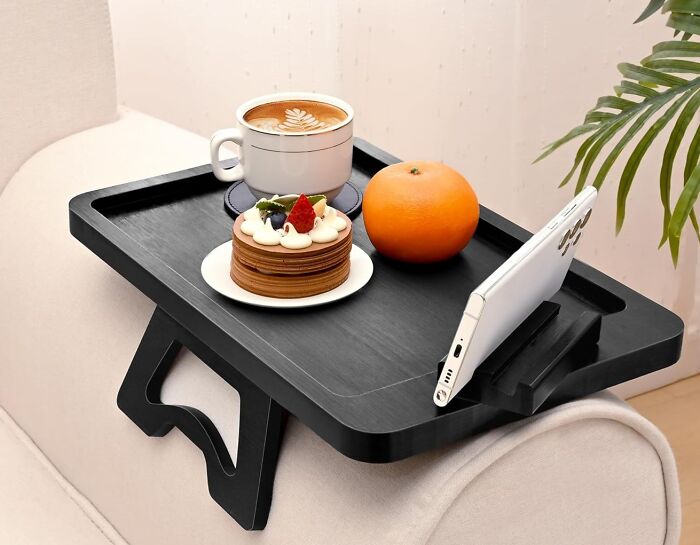 Cozy products for relaxing: tray with coffee, orange, pastry, and phone holder on a sofa arm.