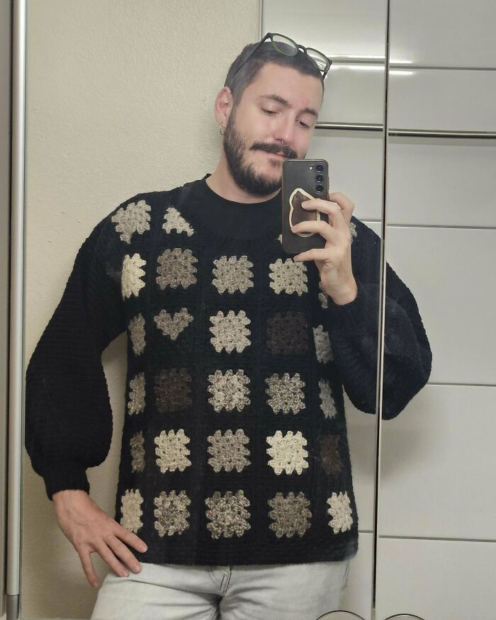 Man taking mirror selfie wearing a handmade crochet sweater with beige and brown square patterns.