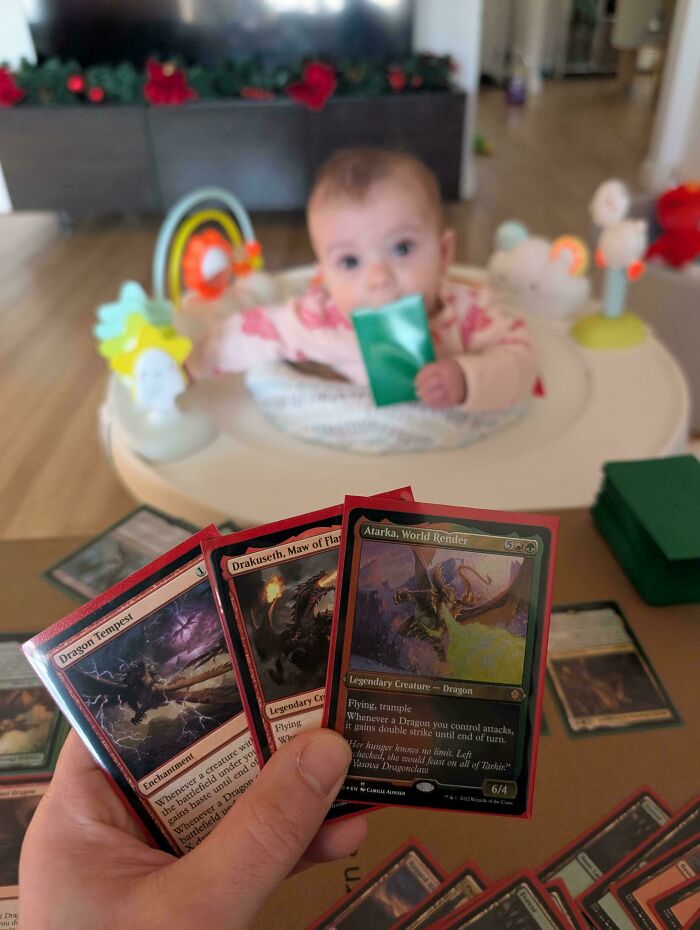 Dad holding Magic: The Gathering cards while baby plays in the background, capturing the rewarding moments of fatherhood.