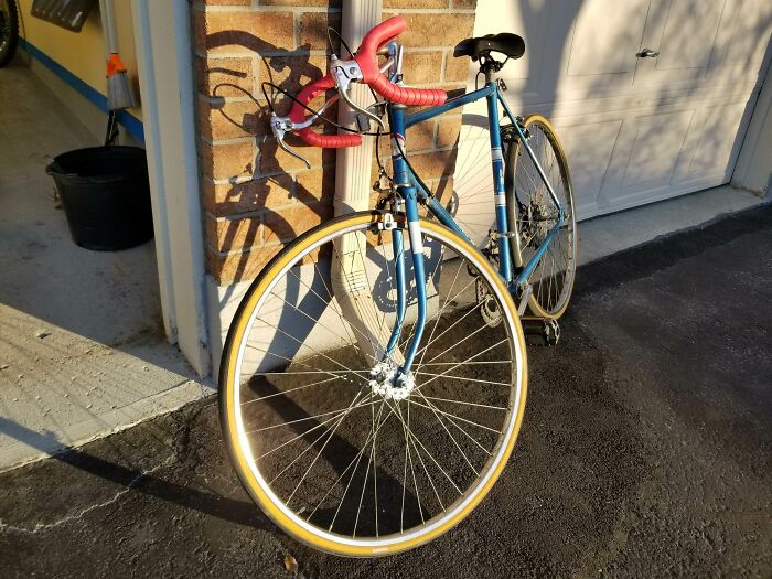 Vintage blue road bike with red handlebars, evoking 80s nostalgia, leaned against a brick wall in sunlight.