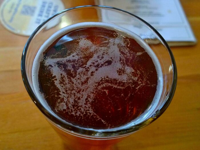 Pareidolia in beer foam, resembling a face on a glass of amber ale on a wooden table.