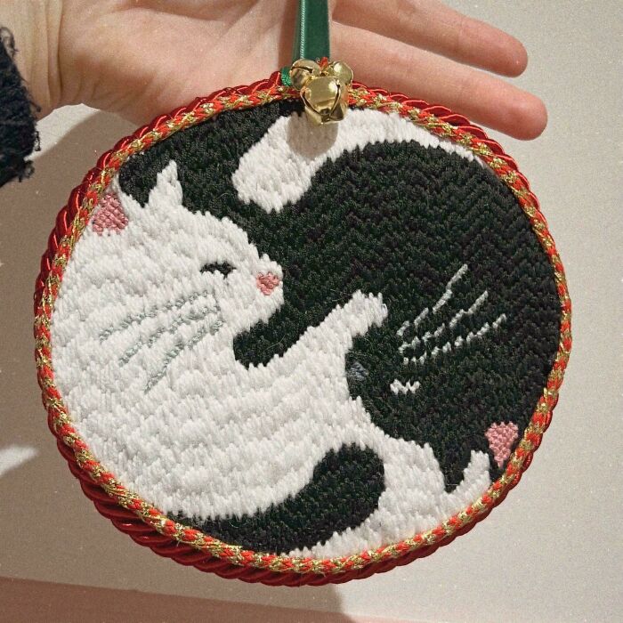 Hand holding a circular embroidery design featuring two intertwined cats in black and white, detailed stitch work visible.