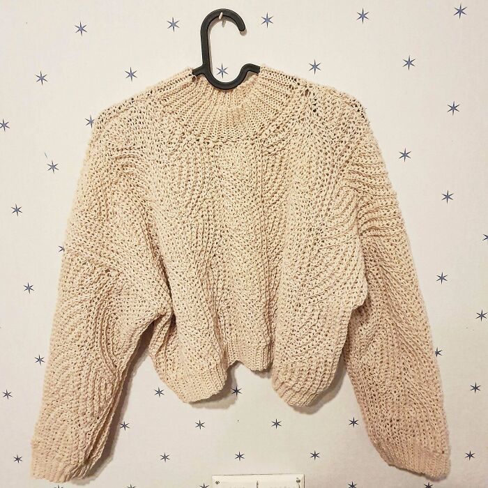 Beige chunky crochet sweater hanging on a black hanger against a white wall with small star patterns.
