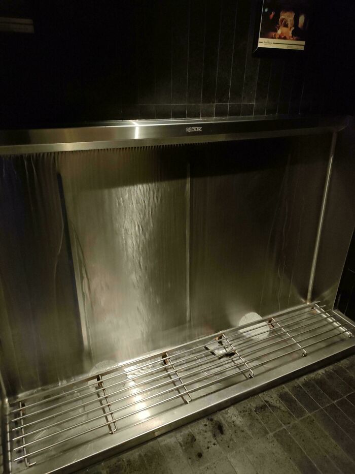Stainless steel urinal with water flow in an Australian restroom, featuring a dark tiled background.