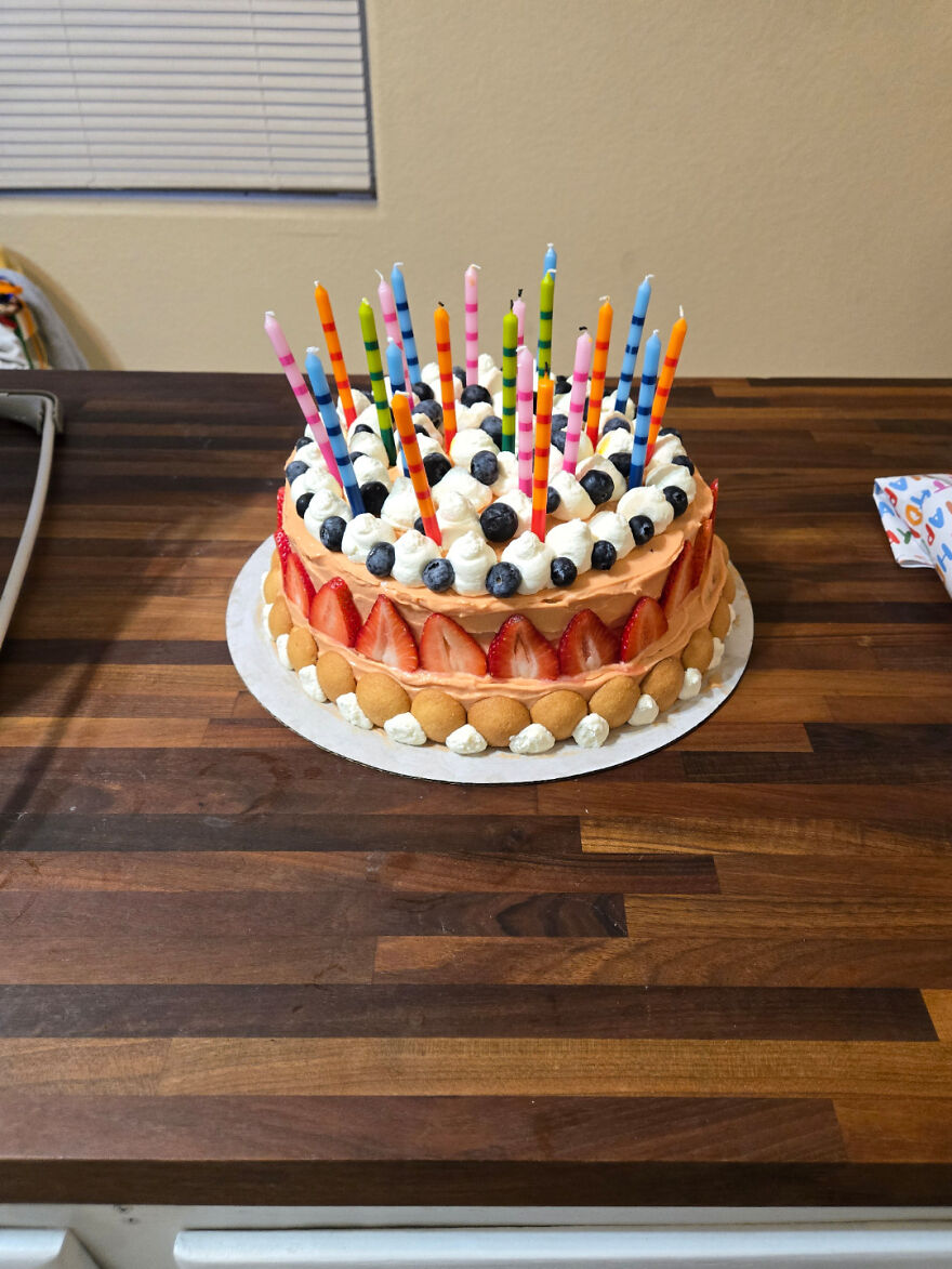 Pastel casero decorado con fresas, ar&aacute;ndanos, crema y velas de colores sobre una mesa de madera oscura.
