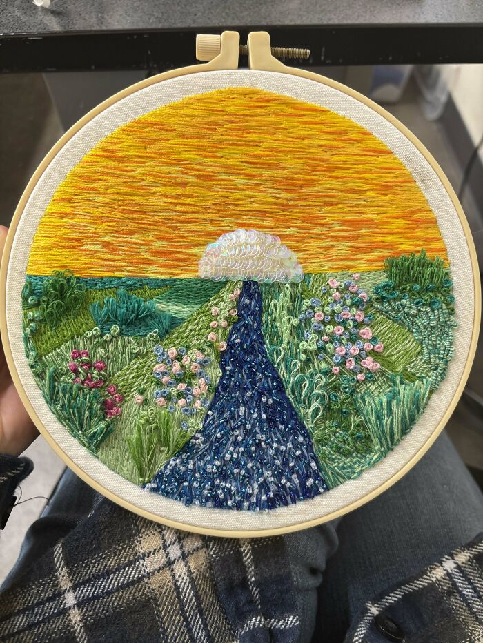 Hand holding embroidery hoop with a vibrant landscape design featuring a sunset, river, and colorful flowers stitched in detail.
