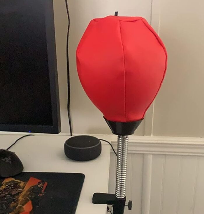 Red desktop punching ball on spring stand next to computer monitor and smart speaker for desk joy.