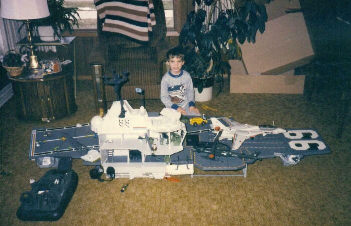 Child playing with large aircraft carrier toy in 80s-style living room, capturing 80s nostalgia.