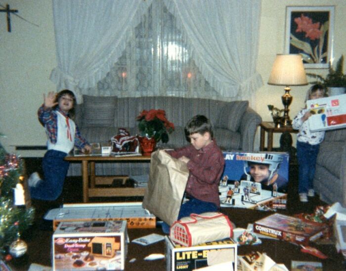 Children in a living room opening Christmas gifts, surrounded by 80s toys like Lite-Brite and Easy-Bake Oven.