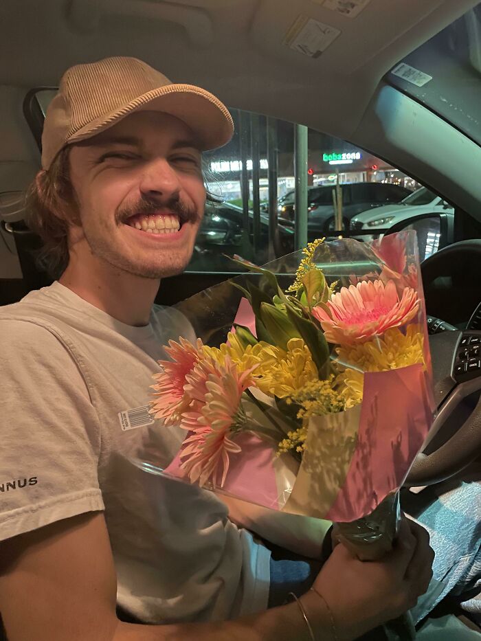 Man smiling in a car, holding colorful flowers, depicting a funny wholesome anniversary surprise.