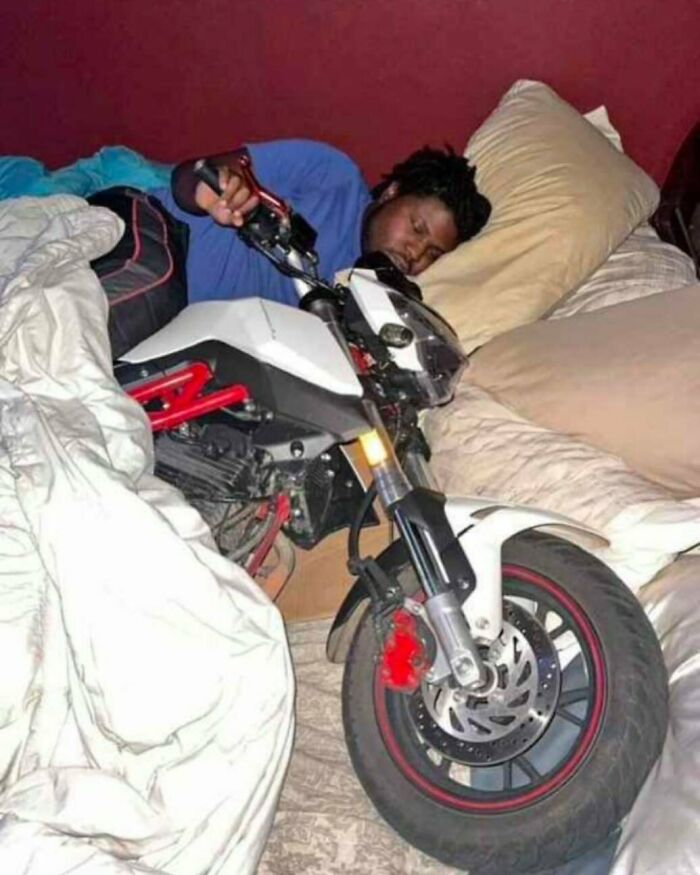 Man lying in bed next to a motorcycle, showcasing a weird side of the internet with unusual sleeping arrangements.