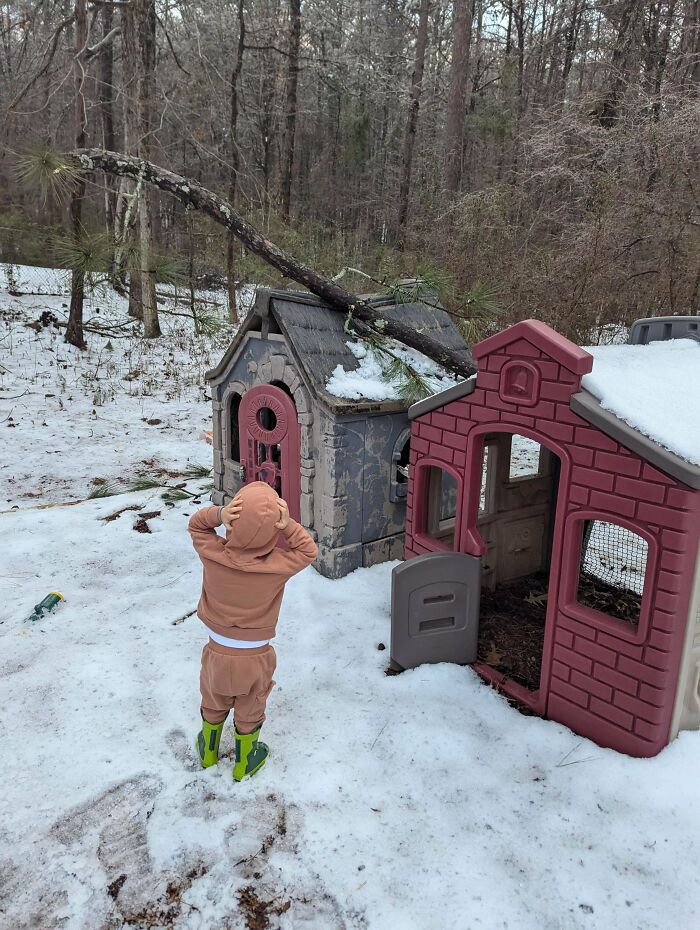 Child in a tan outfit and green boots looking at snow-covered playhouses in a forest.