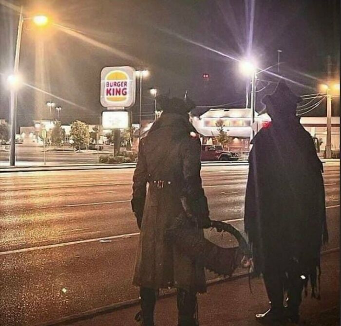 Two people dressed in dark costumes standing on a street near a Burger King sign at night, weird side of the internet.