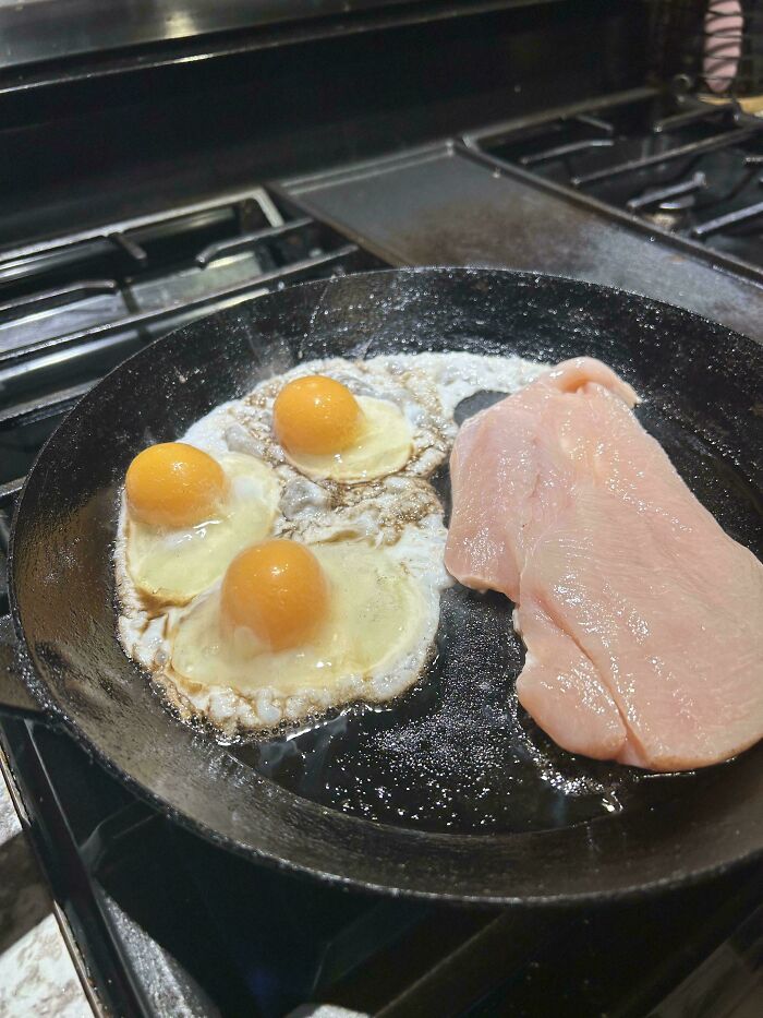 Three oddly shaped fried eggs and a raw chicken breast cooking in a pan, illustrating dishes that are just stupid.