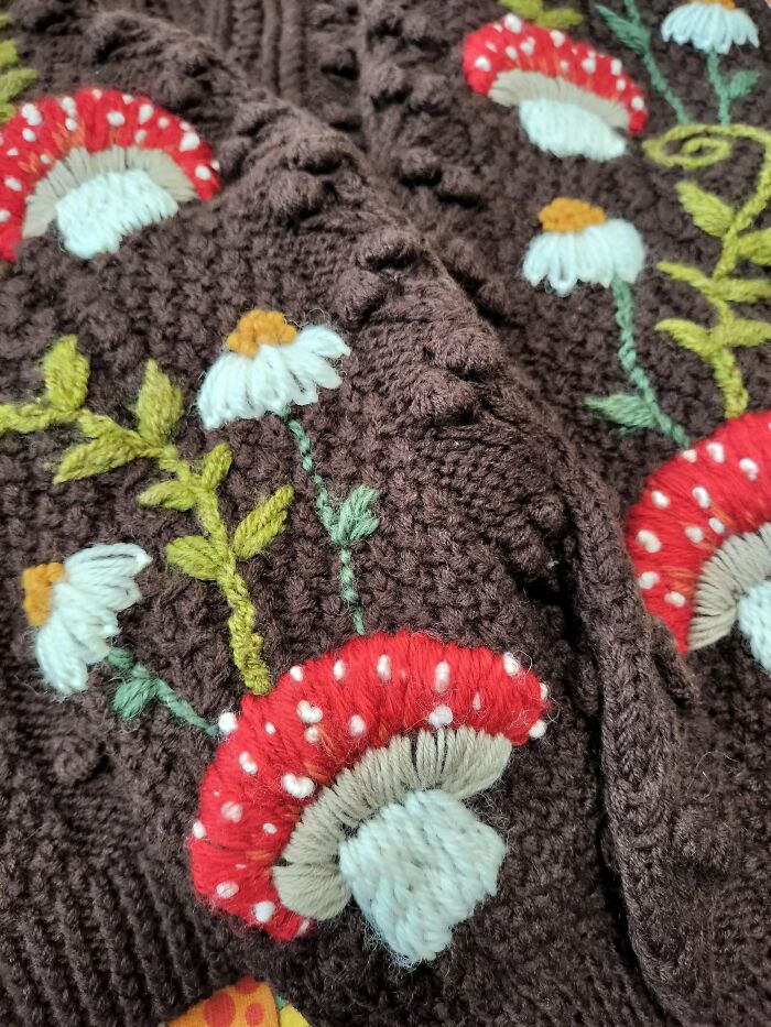Close-up of beautiful embroidery designs with red mushrooms and white flowers stitched on brown knitted fabric.
