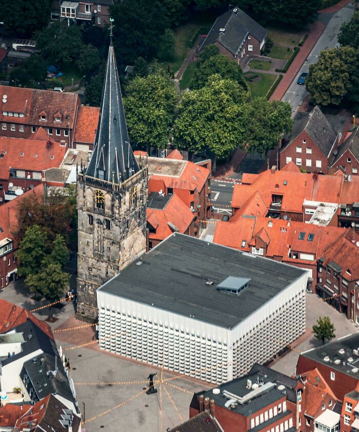 Urban hell: Aerial view of a stark modern building juxtaposed with historic brick rooftops and a church tower.