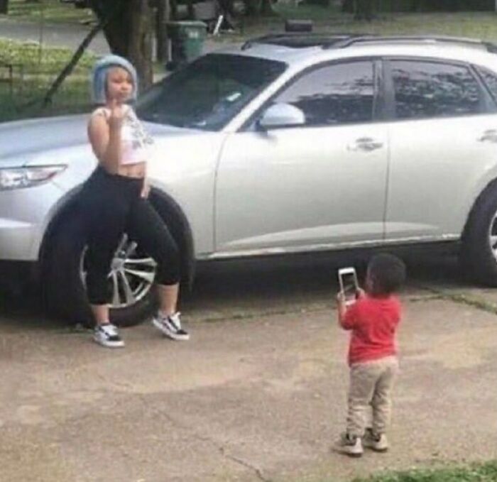 Child taking a photo of woman posing with car wheel in a humorous scene from the weird side of the internet.