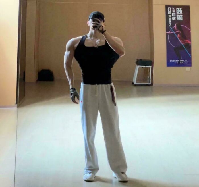 Man posing in gym mirror, wearing black tank top, white pants; strong example of Photoshop airbrush.