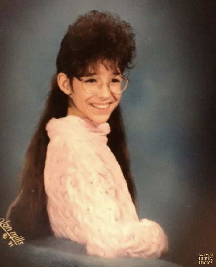 Girl with large glasses and voluminous hairstyle smiling in an awkward family photo with a pink sweater.