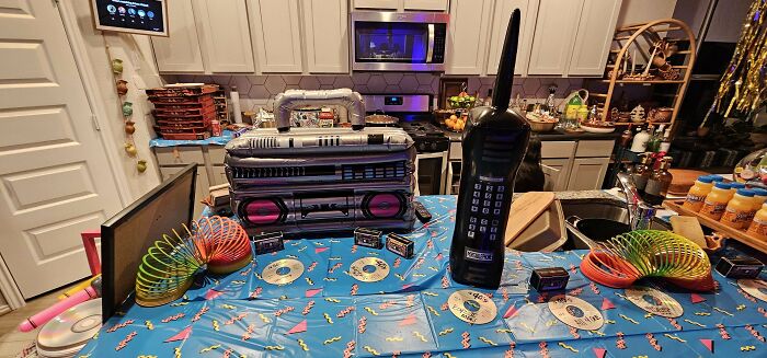 '90s themed kitchen setup with inflatable boombox, giant cellphone, CDs, slinkies, and VHS tapes on a decorated table.