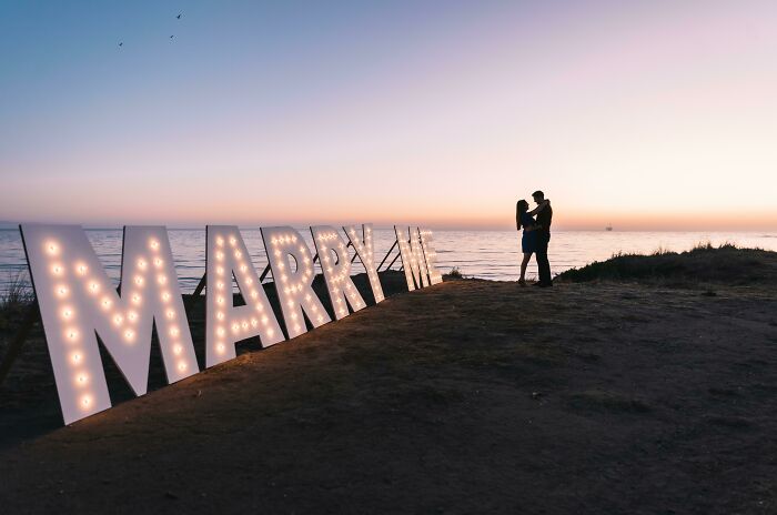 Couple embracing at sunset by "Marry Me" lights, symbolizing long-distance love.