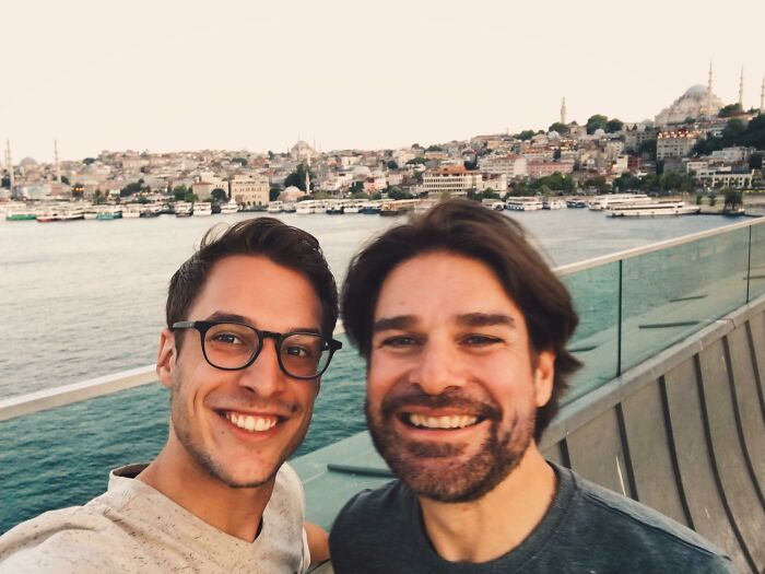 Two men smiling by a waterfront, capturing a long-distance love moment.