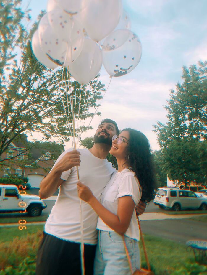 A couple looks up at balloons, symbolizing long-distance love enduring over time, surrounded by trees and cars.