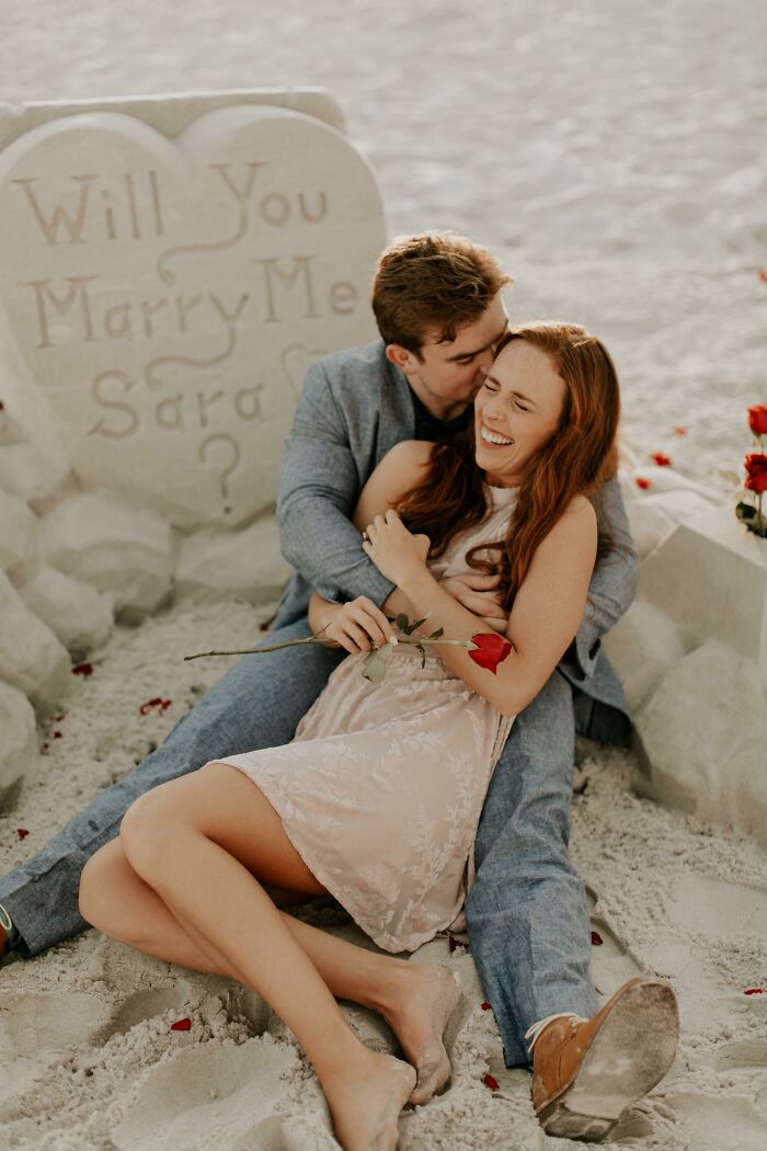 Couple celebrating engagement on a beach, surrounded by roses; heart-shaped sand sculpture with the proposal message.