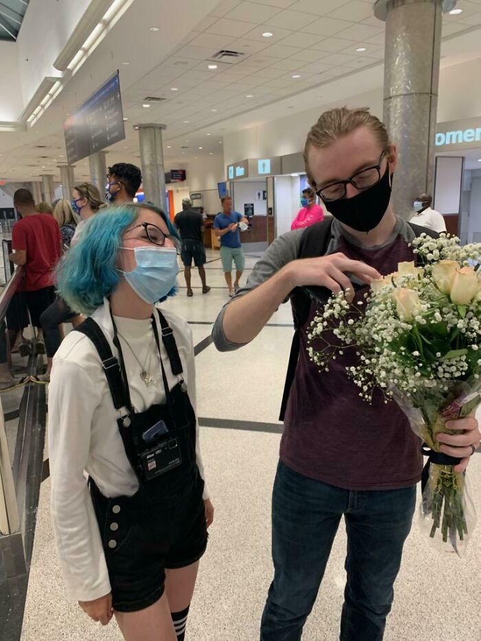A couple with face masks smiles in an airport, holding a bouquet, symbolizing long-distance love.