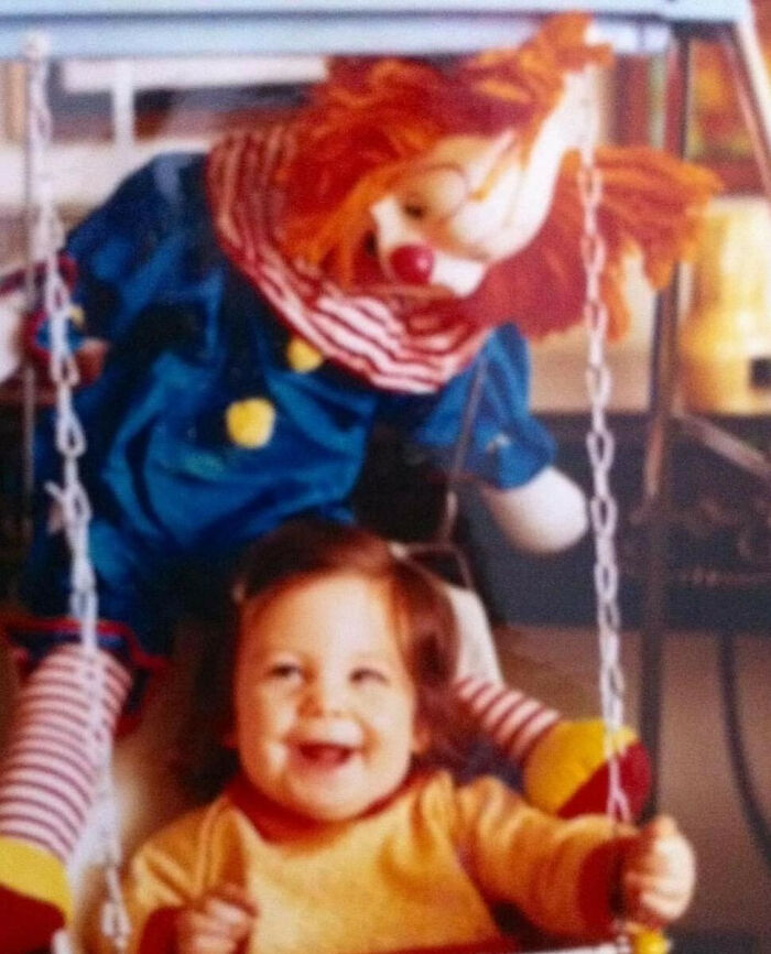 Toddler smiling on swing with a large clown doll in the background in an awkward family photo sharing a sweet moment.