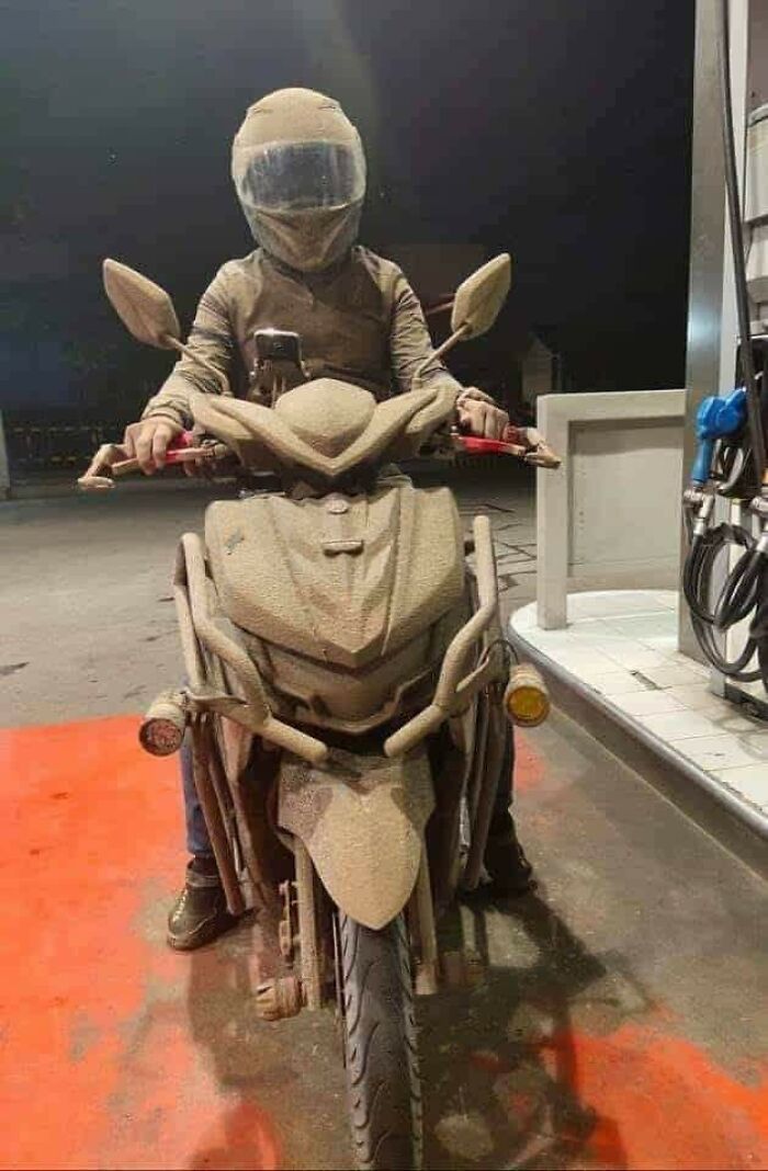 Motorcyclist covered in ash showcases nature disaster resilience at a gas station.