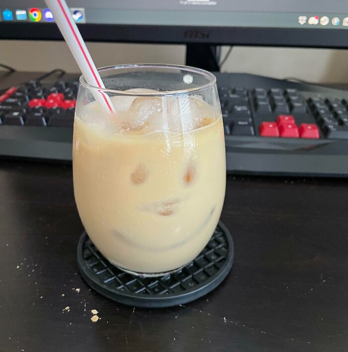 A glass of iced coffee with a smiling face illusion, illustrating pareidolia, on a desk with a computer in the background.