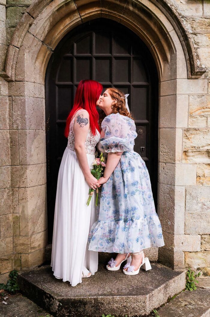 Two women smiling and kissing, wearing elegant dresses, in front of an arched doorway, symbolizing love wins.