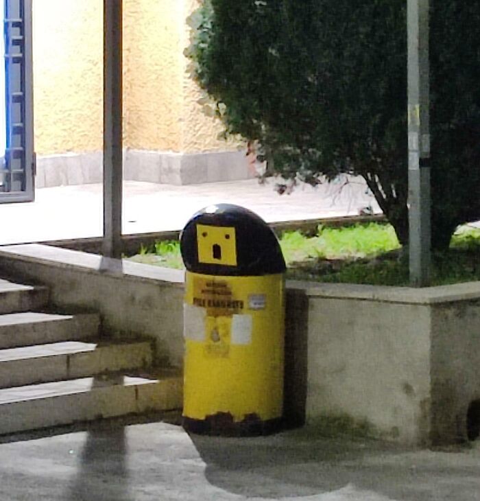 Yellow trash bin resembling a surprised face, demonstrating pareidolia near a staircase and greenery.
