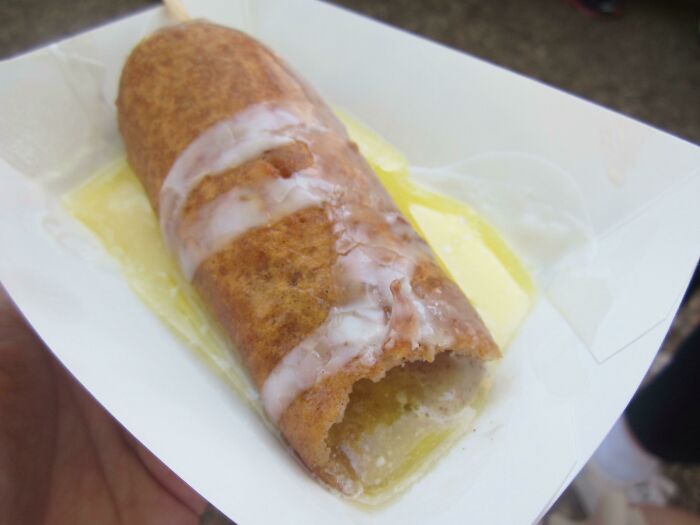 A deep-fried cinnamon roll on a stick, drizzled with icing, showcasing peculiar culinary customs from different countries.